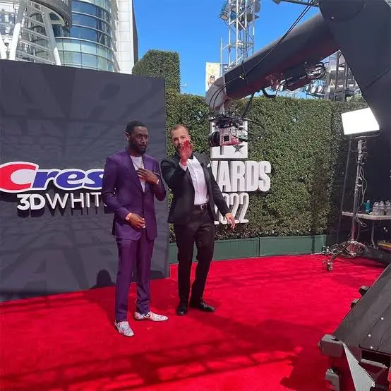 Two men posing on a red carpet at a branded event with a robotic camera arm above them